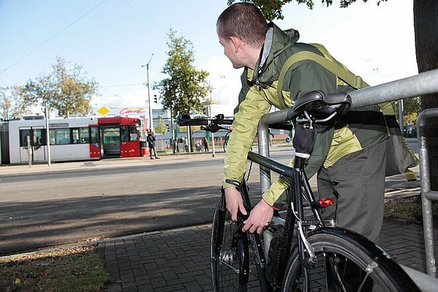 Umfrage Umstieg Rad Mann schließt Fahrrad vom Fahrradbügel ab und blickt auf Straßenbahn im Hintergrund