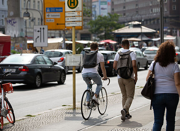Eine Straßenszene: Ein Fahrradfahrer und zwei Fußgänger teilen sich einen gepflasterten Gehweg. Im Hintergrund parken Autos am Straßenrand.