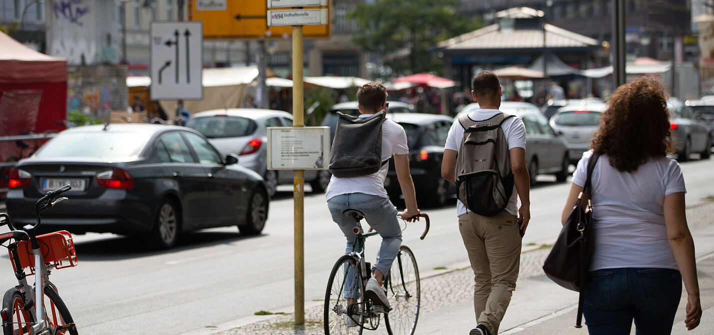 Geteilter Verkehrsraum in der Stadt Eine Straßenszene: Ein Fahrradfahrer und zwei Fußgänger teilen sich einen gepflasterten Gehweg. Im Hintergrund parken Autos am Straßenrand.