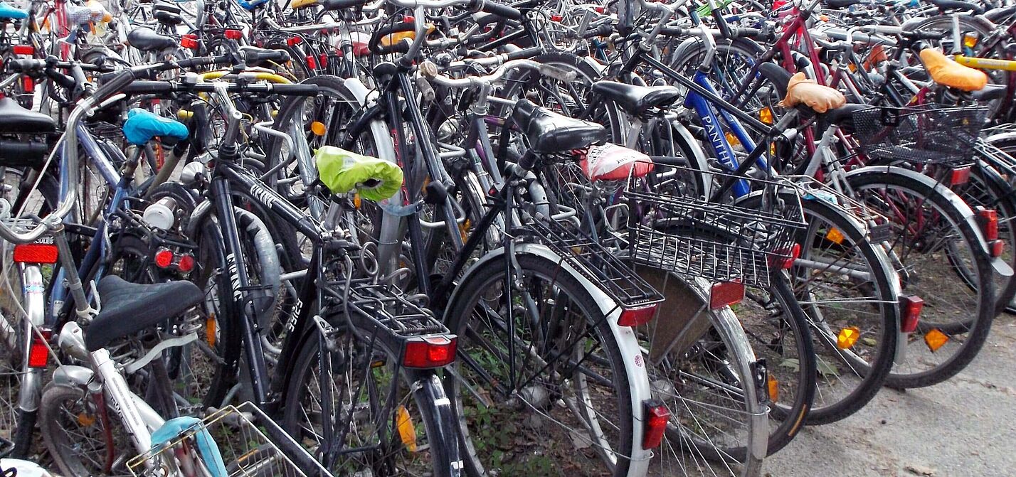 Fahrradparkplatz ohne Abstellanlagen Hunderte von Fahrrädern verschiedener Farben und Modelle stehen dicht gedrängt und ungeordnet auf einer befestigten Fläche; die Räder sind ineinander verschachtelt, mit Körben, Taschen und bunten Sattelbezügen ausgestattet, ohne erkennbare Parkordnung oder Abstellvorrichtungen.